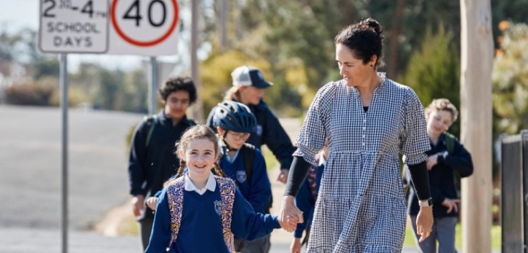 Female guardian holds the hand of young female student as they walk and smile, leading a line of students on a pavement in front of a school zone sign.